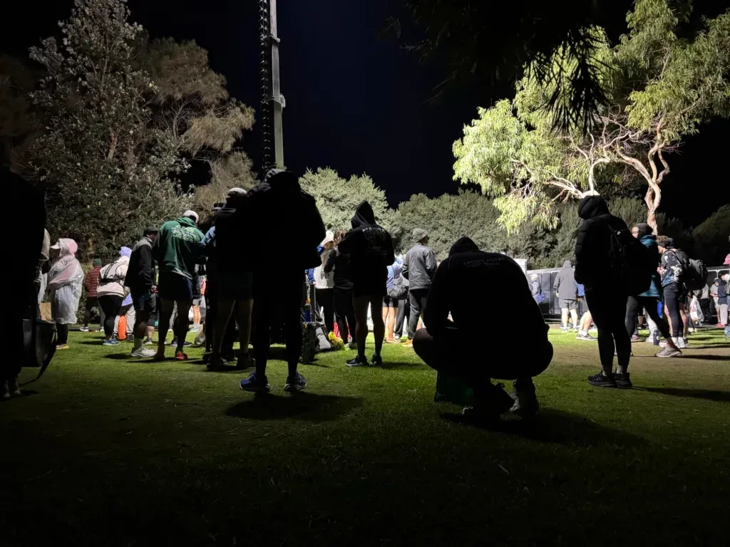 Runners in hoodies and jackets grouped on floodlit grass in the dark, one crouched down tying a shoe