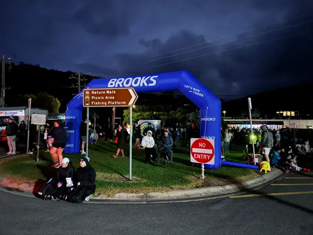 Blue inflatable Brooks arch over the road before dawn, two people in beanies huddled on the kerb below it