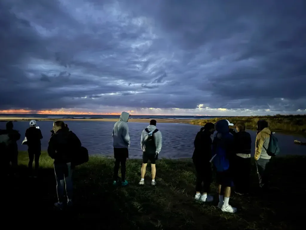 Runners in hoodies standing along the grass above a lagoon, watching orange light break under thick cloud