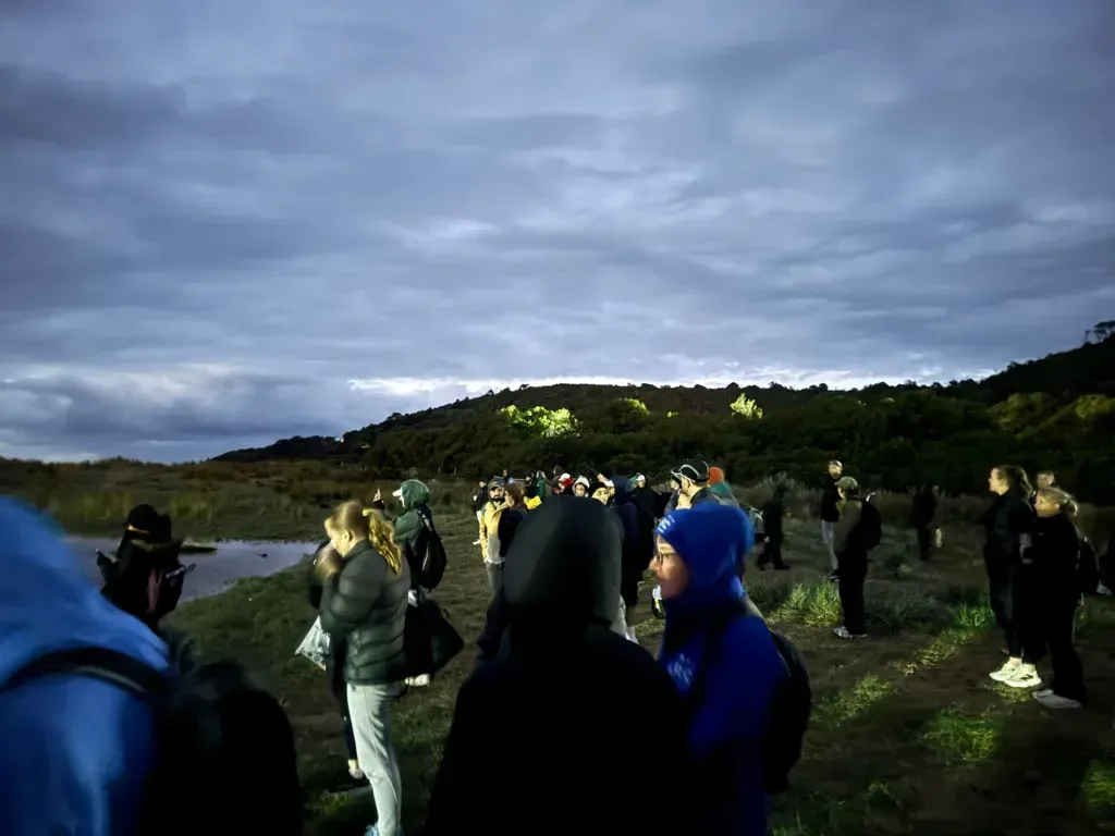 Crowd in beanies and puffer jackets waiting on the grass in blue half-light, bush-covered hills behind