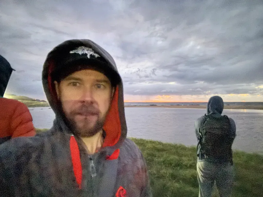Kyle in a hood and cap taking a selfie by the water, another runner with a backpack watching the sunrise