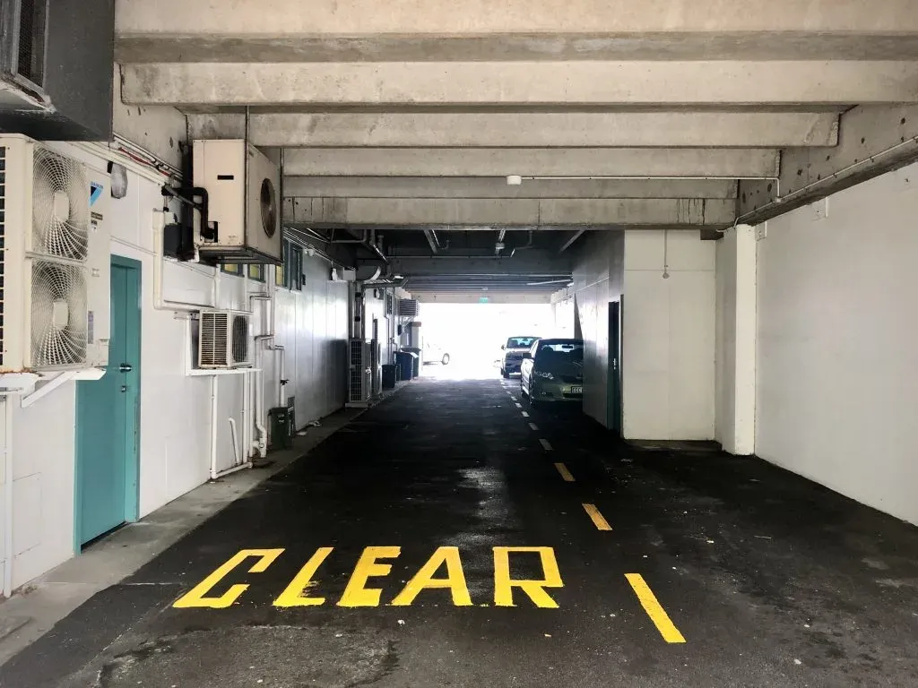 Car park driveway with CLEAR painted in yellow on the asphalt, air-con units along the wall, cars at the bright exit