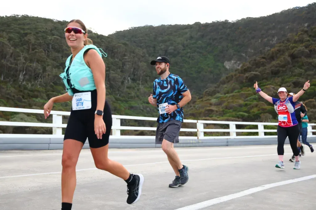 Kyle jogging across a bridge in a blue patterned shirt, other runners ahead and behind, bush hills beyond